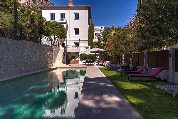A large white villa with a reflecting swimming pool, a terraced garden with lounge chairs, and lush trees under a bright blue sky.