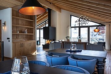 Modern mountain chalet interior with wooden ceilings, cozy blue chairs, a burning fireplace, and snowy winter views through large windows.
