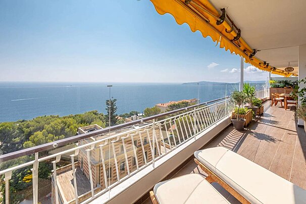 A spacious balcony with lounge chairs, a dining area, and a yellow awning overlooks a stunning panoramic ocean and coastline under a clear blue sky.