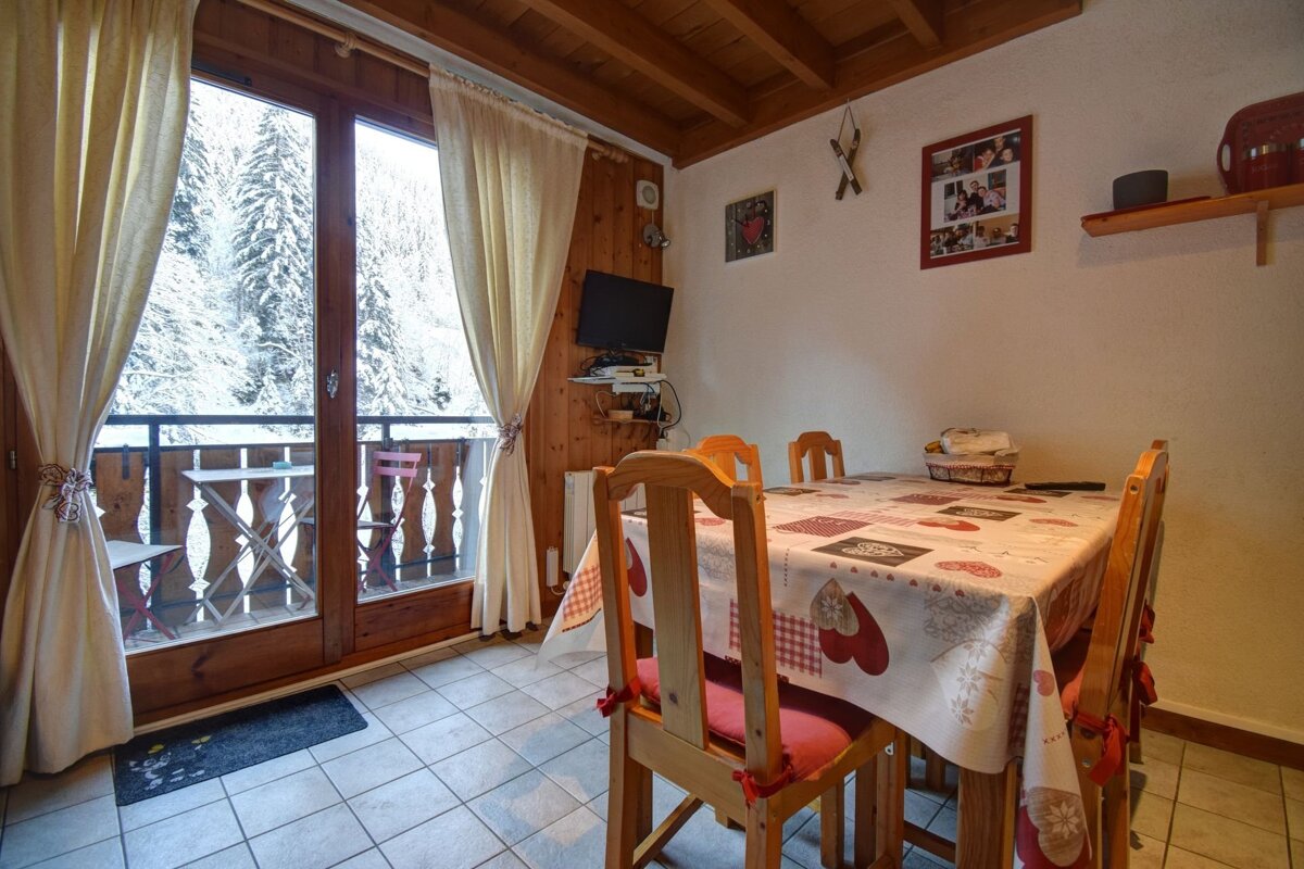 A cozy dining room with a table, chairs, and a balcony offering a snowy forest view. Wooden ceiling, small TV, and wall decorations.