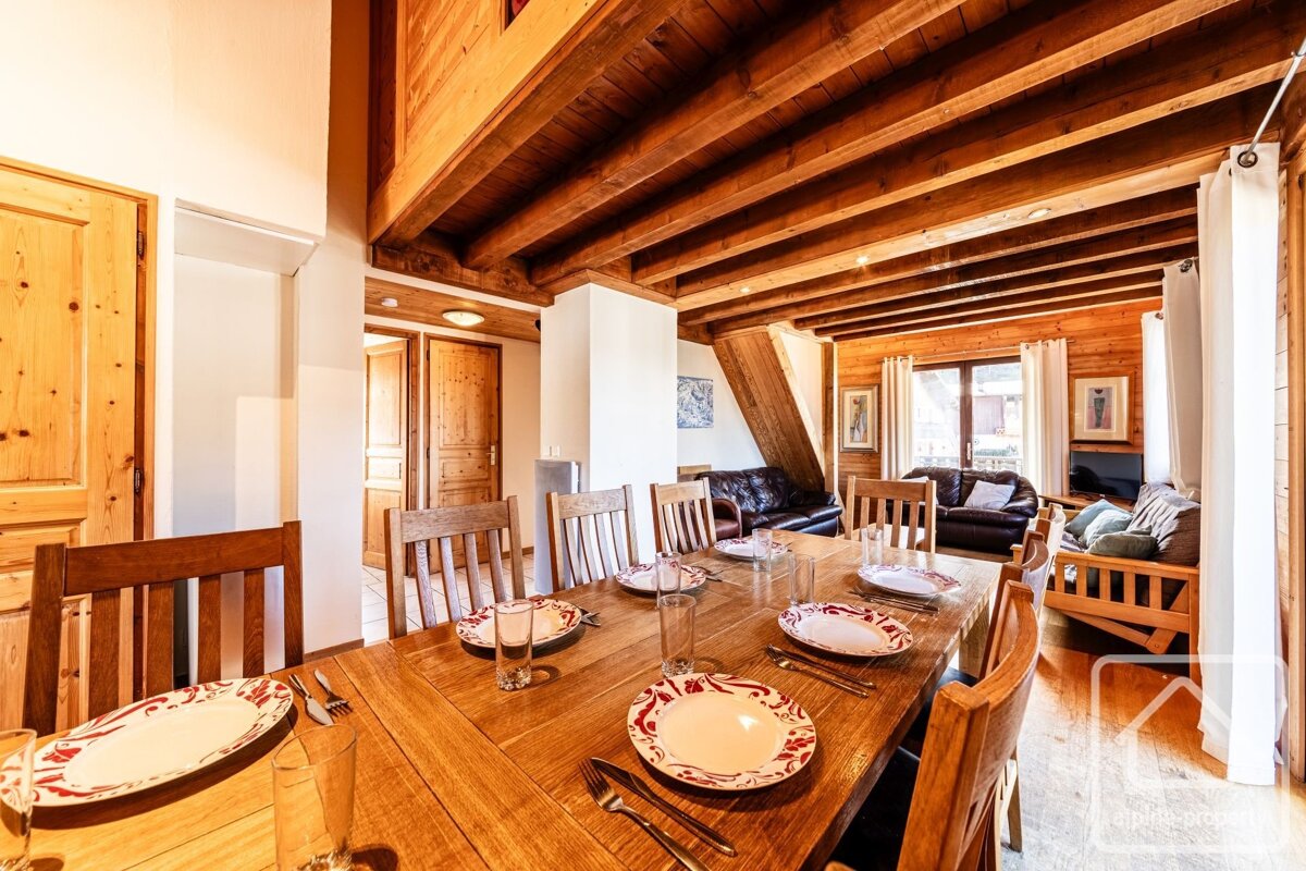 A warm, wood-filled chalet interior: a dining room with a long table, set with plates, open to a living area with leather sofas, all under exposed beams.