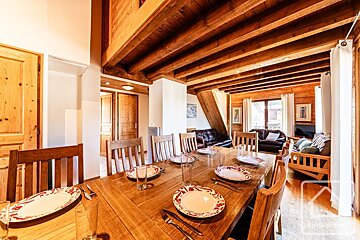 A warm, wood-filled chalet interior: a dining room with a long table, set with plates, open to a living area with leather sofas, all under exposed beams.