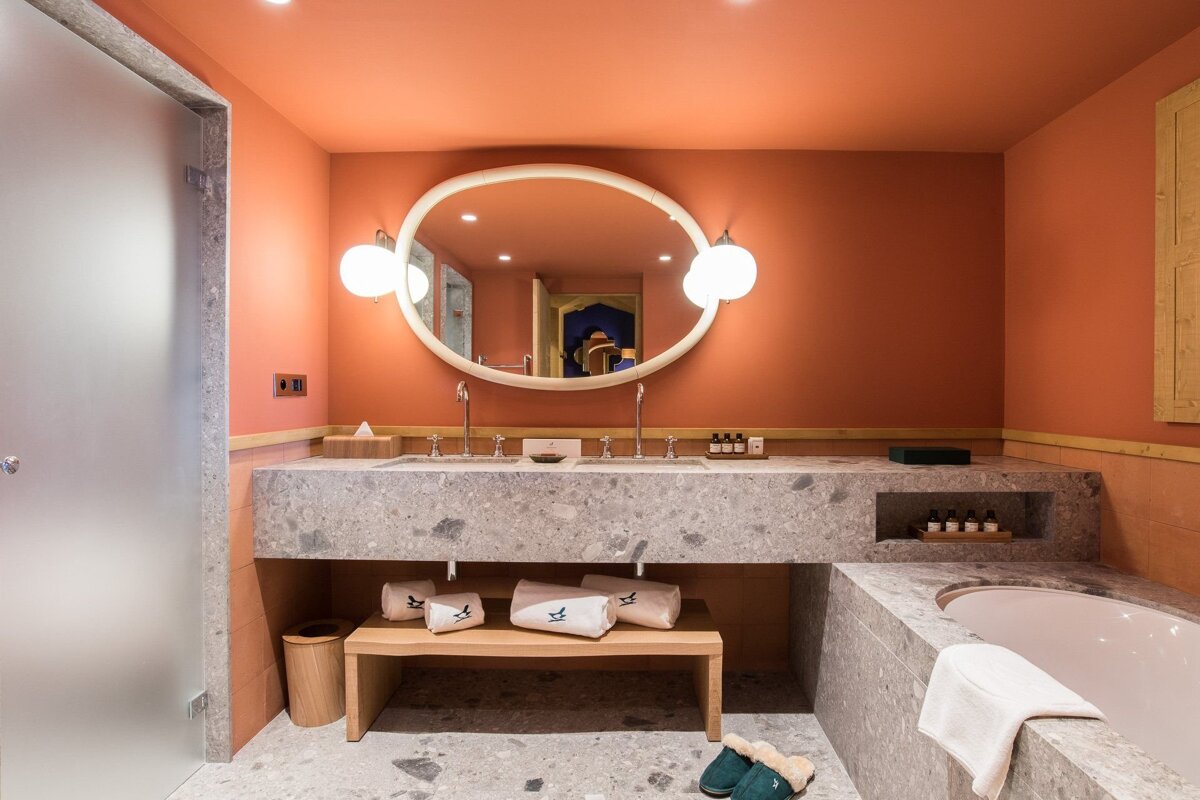 A chic bathroom with terracotta walls, grey speckled stone double vanity and tub, an oval mirror, and slippers. Modern and inviting design.