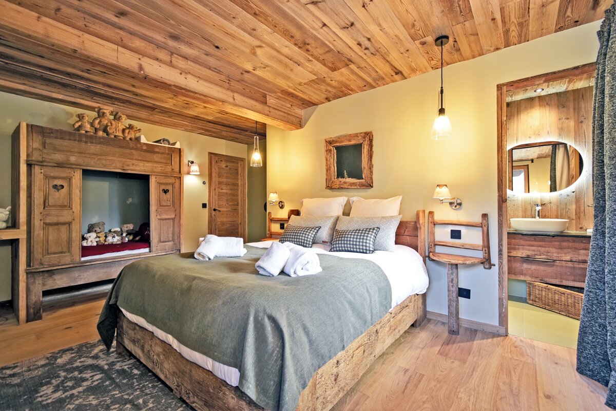 Cozy, rustic bedroom with a wooden plank ceiling, a large bed, and a built-in sleeping nook adorned with teddy bears. An open sink area is visible.