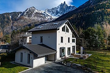 A sleek modern white house with a dark roof is set against a backdrop of majestic snow-capped mountains and an autumnal forest in a sunny valley.