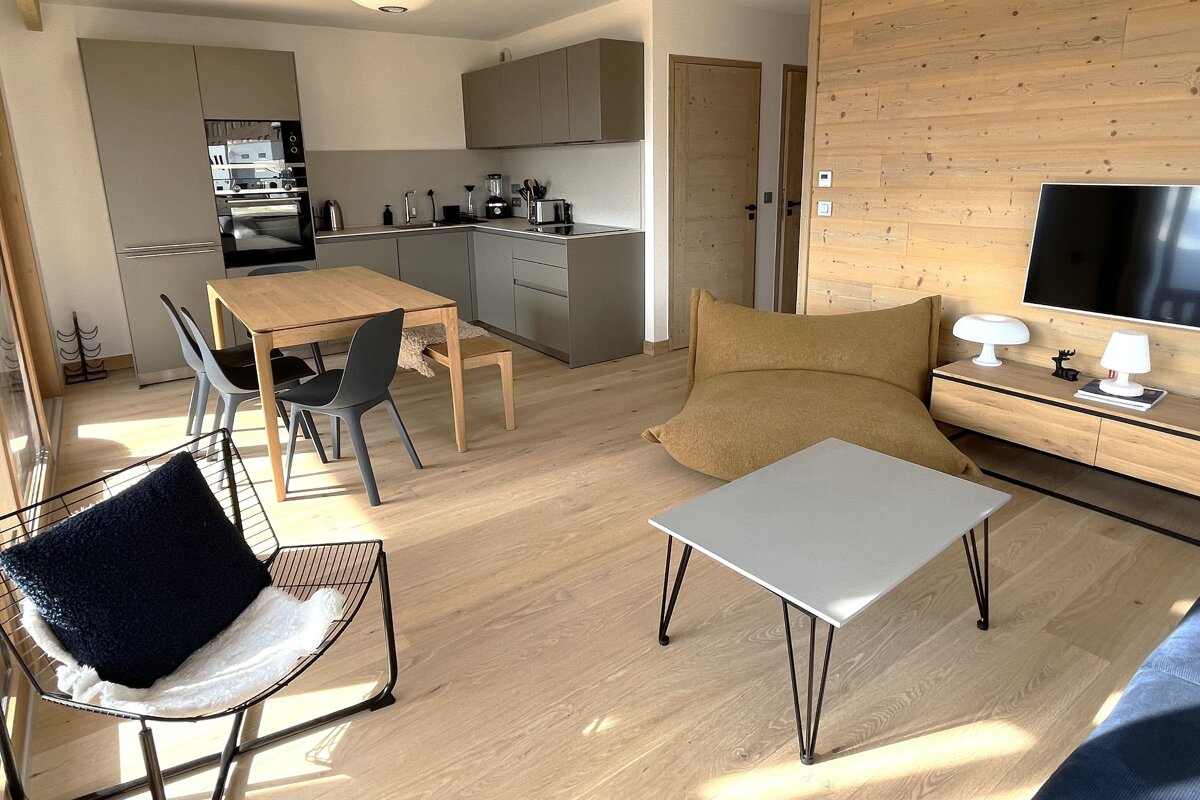 A bright, modern open-plan room: grey kitchen, wood dining table, living area with yellow beanbag, coffee table, TV, and wood accent wall.