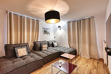 Cozy living room with grey modular sofa, light beige curtains with blue accent lighting, wooden floor, and modern light fixtures.