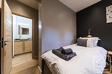 Modern bedroom with a single bed, folded towels, and an open doorway showing an ensuite bathroom with a wooden vanity.