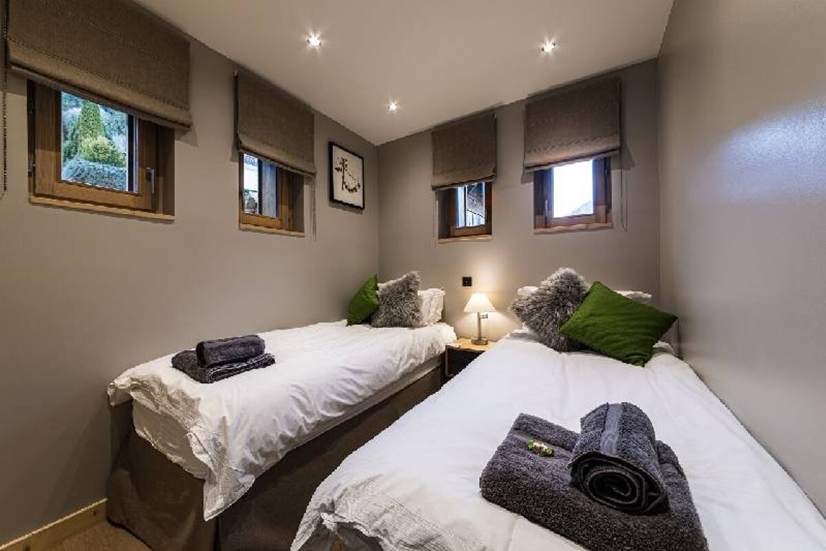 Cozy bedroom with two twin beds featuring white linens, green and grey pillows, folded towels, and small windows with roman shades.