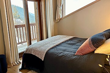 A cozy bedroom with dark bedding, accented by colorful pillows, and a balcony overlooking a winter mountain view.