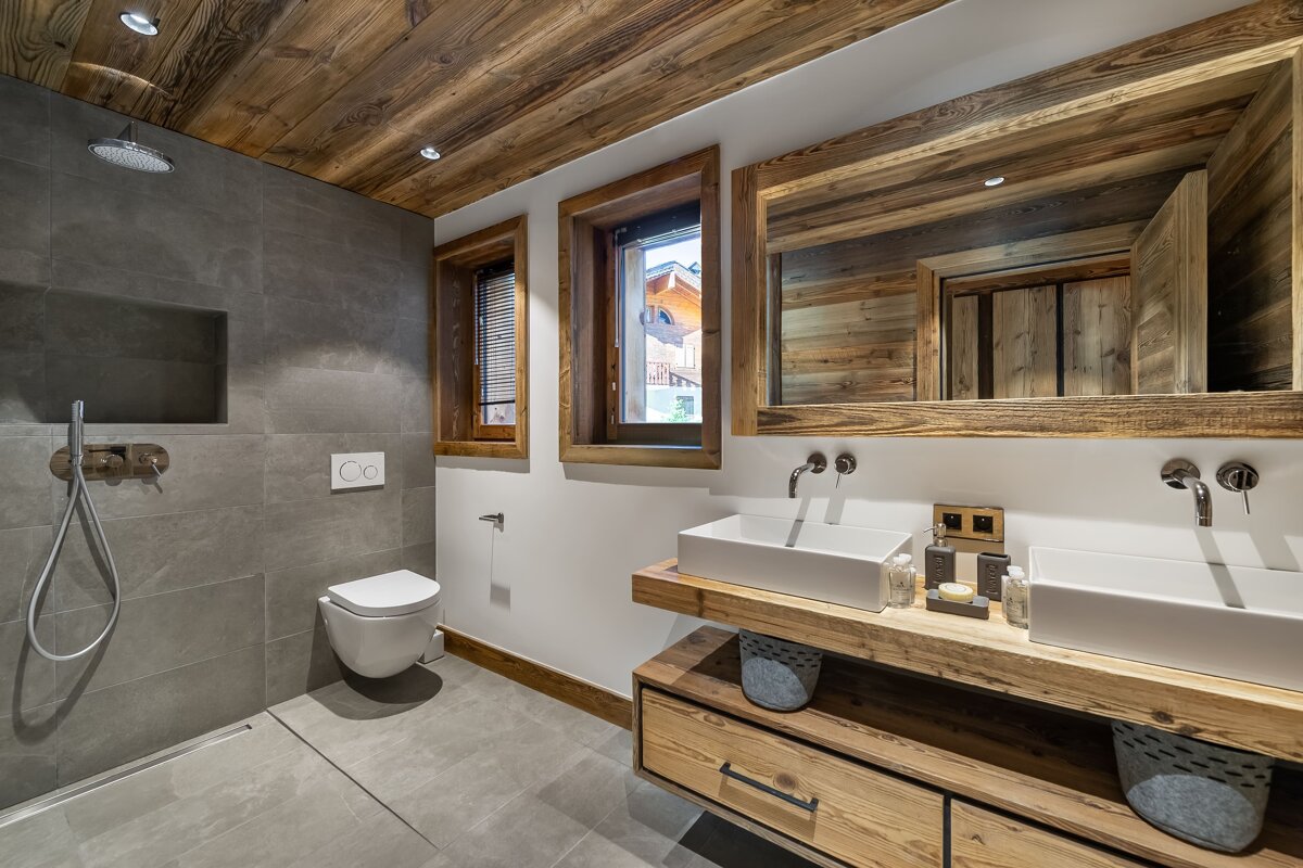 Modern chalet-style bathroom featuring a wooden ceiling, double vanity, wall-mounted toilet, and grey tiled shower.