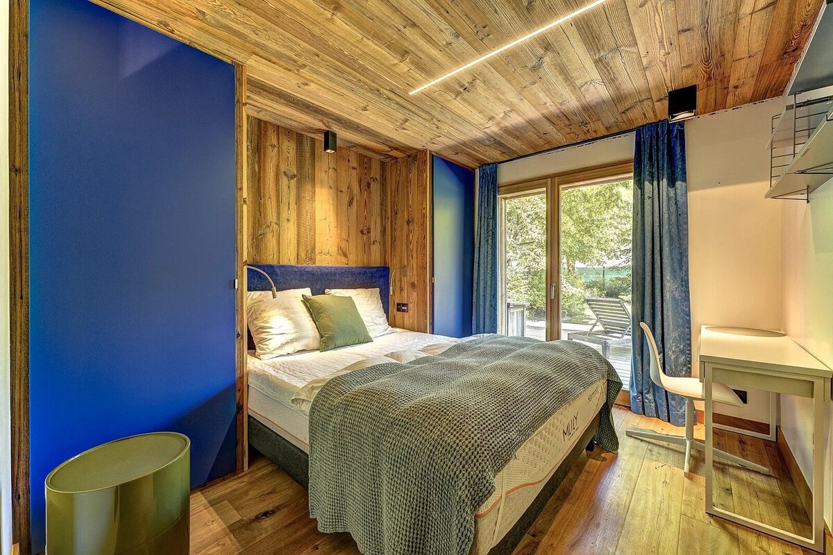 A modern, cozy bedroom with rich wooden walls & ceiling, a royal blue accent wall, a comfortable bed, and a large window to the outdoors.