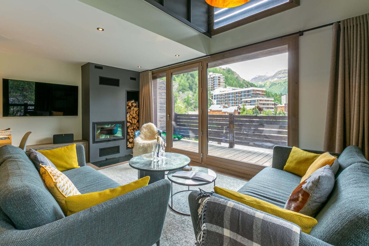 A modern living room with gray sofas, a fireplace, and large windows opening to a mountain town view from a balcony.