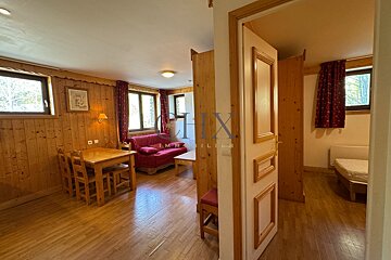 Cozy alpine cabin interior with a wooden dining set, red sofa, wood paneling, and a bedroom visible through an open doorway.