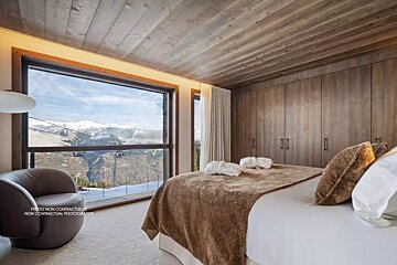 Modern bedroom with a panoramic view of snowy mountains and ski lifts through a large window. Features wooden ceilings, built-ins, and a comfy bed.