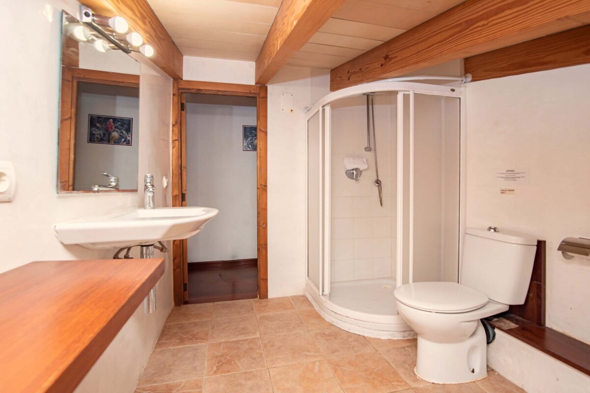 A rustic bathroom featuring a white sink, mirrored vanity, shower cabin, and toilet, with exposed wooden ceiling beams and tiled floor.
