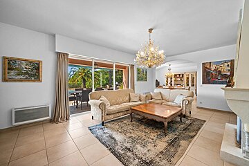 Elegant living room with beige sofas, a wooden coffee table on a rug, a crystal chandelier, and glass doors opening to a garden patio.