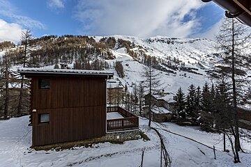 A snow-covered alpine village with wooden chalets and trees, set against a vast snowy mountain range under a partly cloudy blue sky.