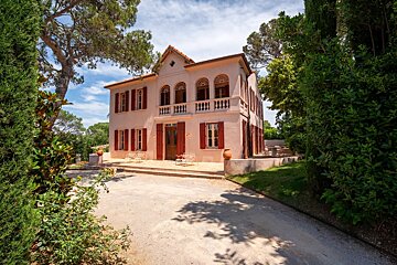 A lovely pink villa with red shutters, a balcony, and arched windows, nestled among lush green trees under a bright blue sky.