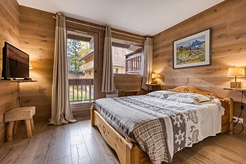 A cozy, wood-paneled bedroom features a wooden bed with patterned bedding, two lamps, and a TV. A balcony door offers a view of other rustic buildings.