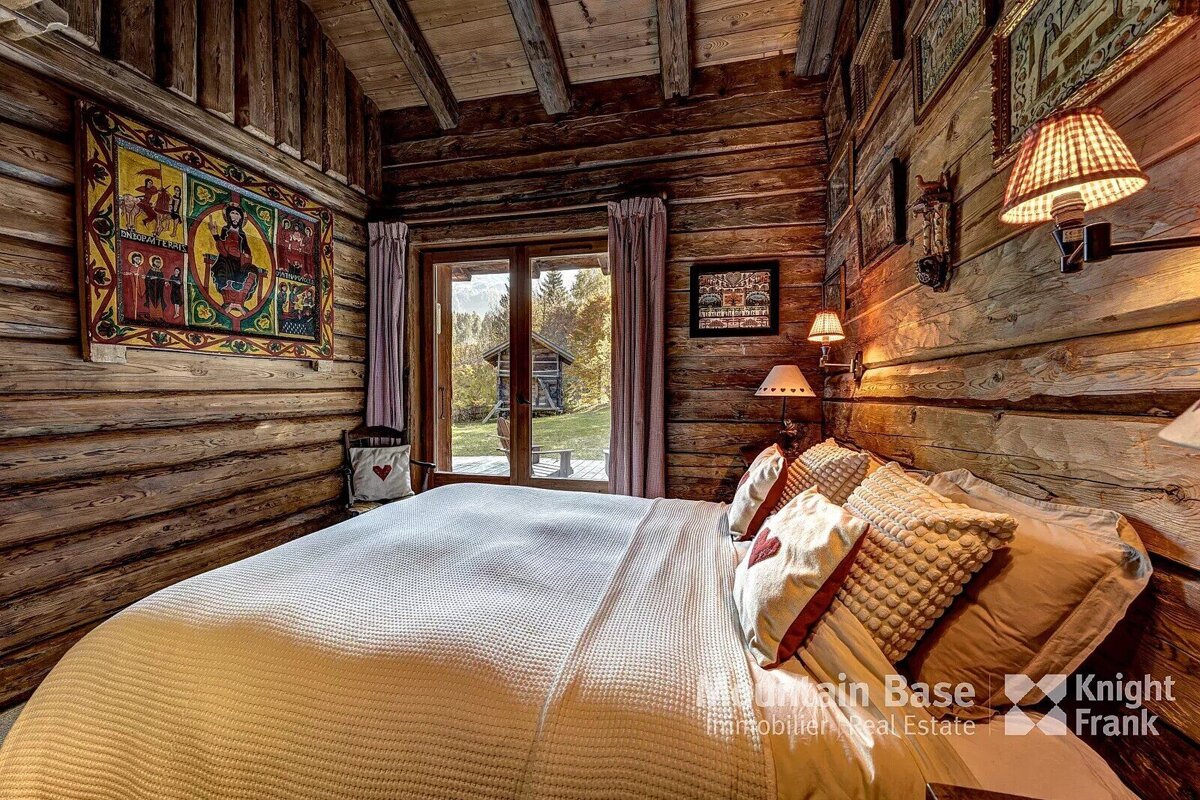 Cozy log cabin bedroom with a large bed, rustic wooden walls, warm lighting, and a window view of another cabin and trees outside.