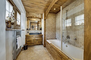 A rustic bathroom with warm wood ceiling, vanity, and tub surround. Features a dark sink, tiled shower/tub, and a towel warmer.