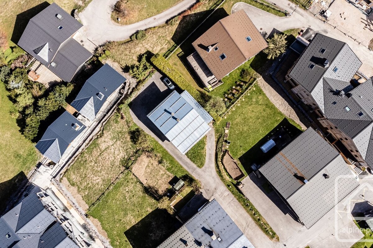 An aerial view shows a cluster of houses with dark, brown, and solar-paneled roofs, surrounded by green lawns and winding roads on a sunny day.