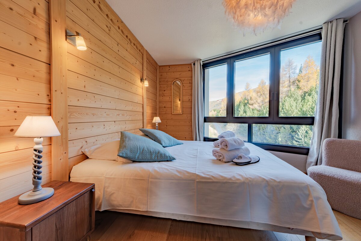 Cozy chalet bedroom with wood-paneled walls, a double bed with blue pillows, and a large window overlooking autumn trees.