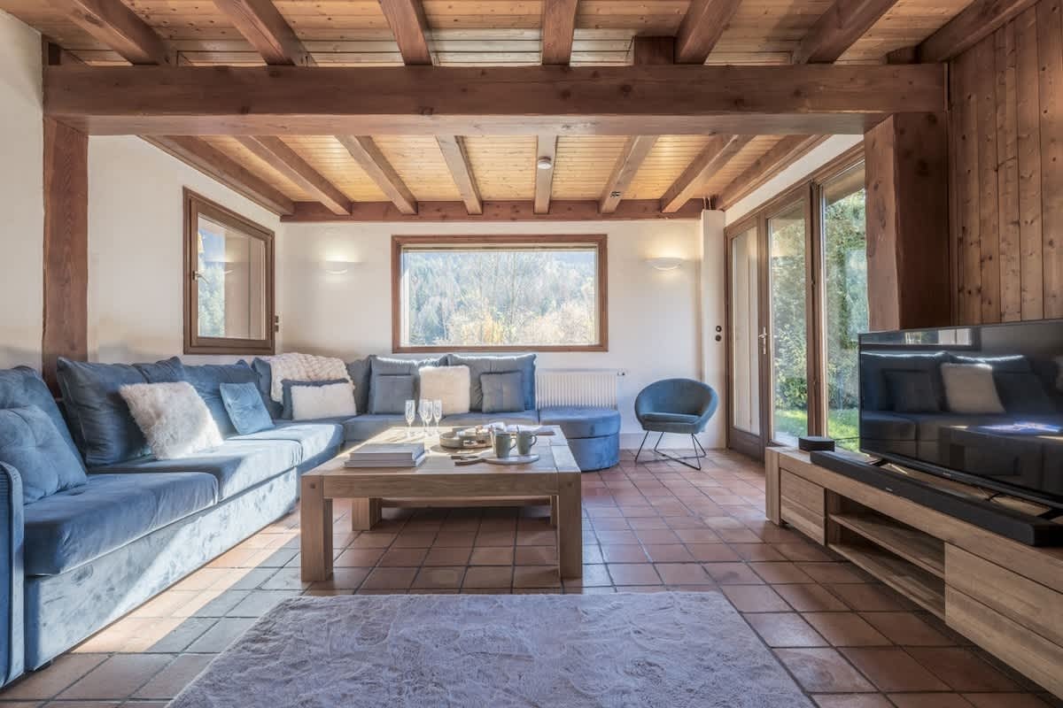 Rustic living room with a large blue sectional sofa, exposed wooden beams, tiled floor, and a TV stand.