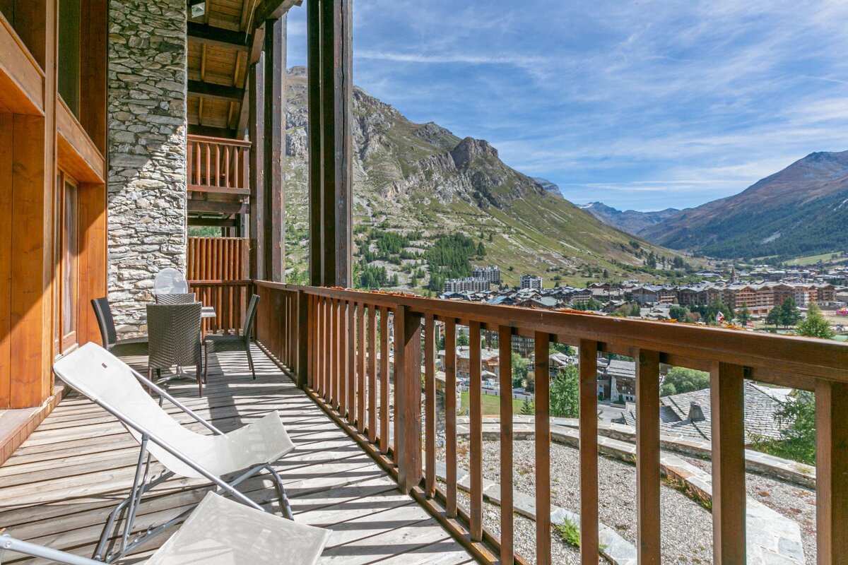 Sunny wooden balcony with chairs and table, offering panoramic views of a mountain village and valley under a blue sky.