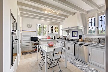 Bright country-style kitchen with white cabinetry, exposed ceiling beams, a small dining table, and tiled flooring.