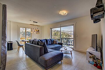 A modern living room featuring a blue sectional sofa, coffee table, TV, and large glass doors overlooking a scenic mountain view.