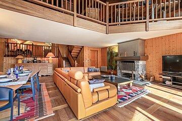 Spacious, multi-level chalet living space with warm wood finishes, leather sofas, a stone fireplace, dining area, and an upper loft.