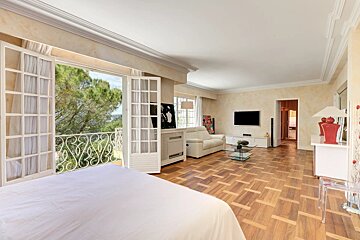 A bright room combining a bedroom and living area, featuring a large balcony with a scenic view of trees, a wooden floor, and a TV setup.