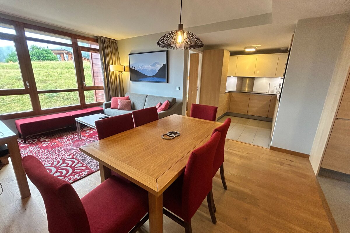 Modern open-plan living/dining/kitchen with red accents, wooden furniture, and large windows looking out to a green, hilly landscape.