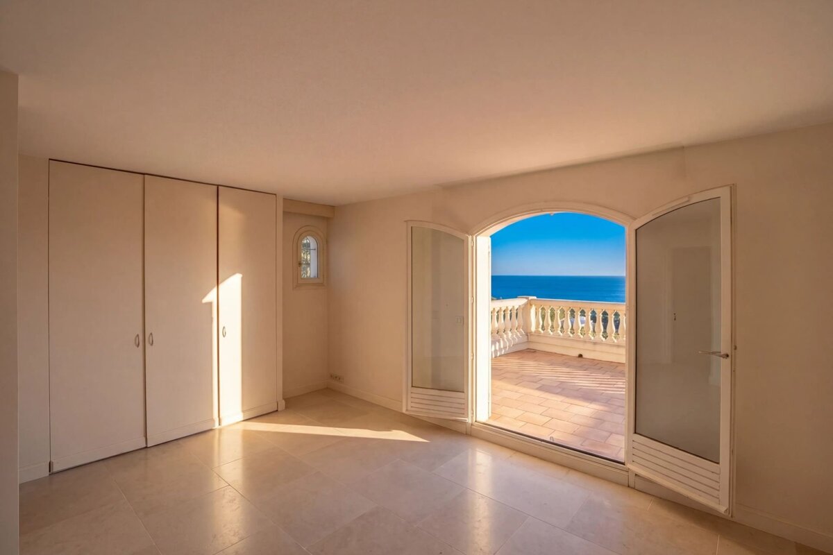 An empty, sunlit room with fitted wardrobes and open arched doors leading to a balcony with sea views.