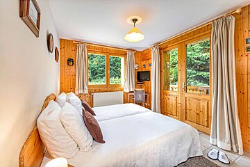 Cozy wood-paneled bedroom with twin beds, a small TV, and large windows/door overlooking a lush green forest, exuding a rustic cabin feel.