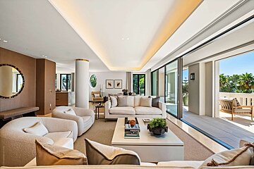 A modern, luxurious living room in neutral tones with large glass doors opening to a sunlit balcony featuring outdoor seating and palm trees.