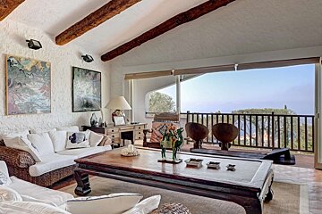 A rustic living room with beamed ceilings, wicker furniture, and a large wooden coffee table, opening to a balcony with a sweeping sea view.