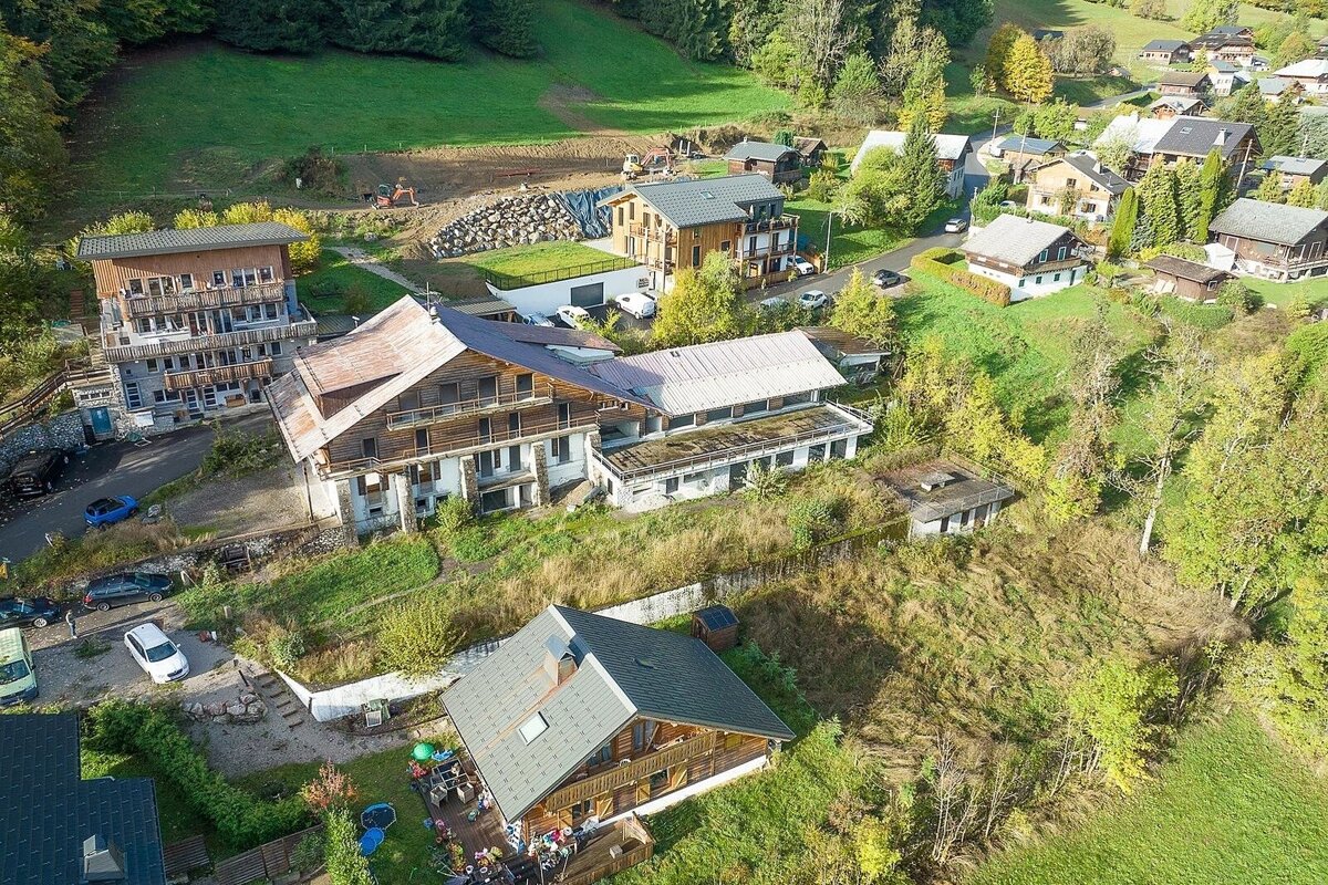 Aerial view of a mountain village: houses built into green, tree-covered slopes, mixing old and new architecture, with visible construction work.