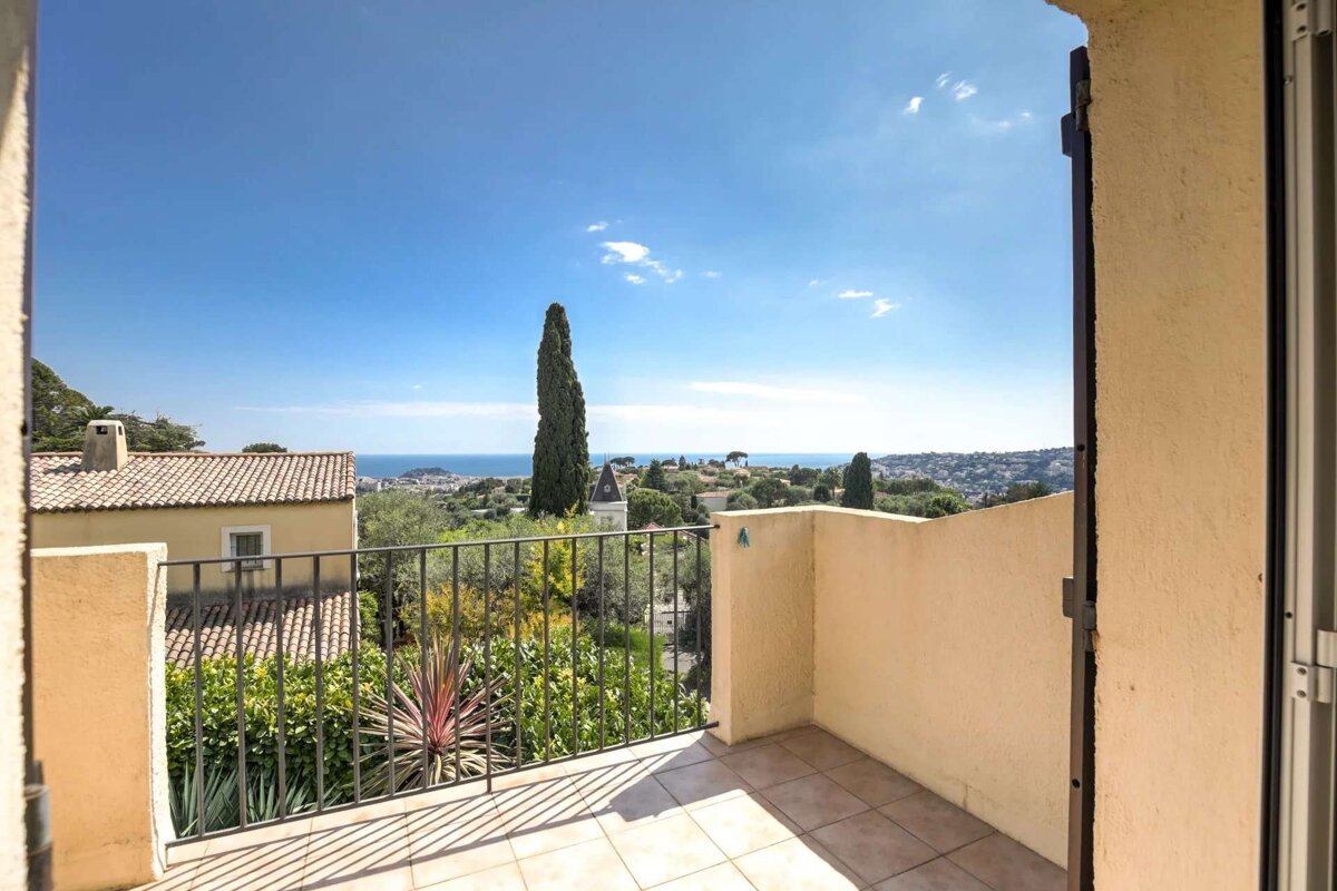 A sunny balcony offers a panoramic view of the blue sea, a coastal town, green hills, and a tall cypress tree under a clear sky.