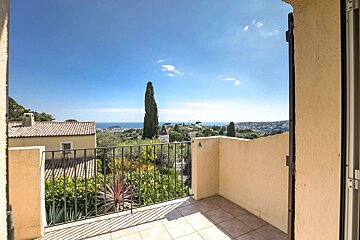 A sunny balcony offers a panoramic view of the blue sea, a coastal town, green hills, and a tall cypress tree under a clear sky.