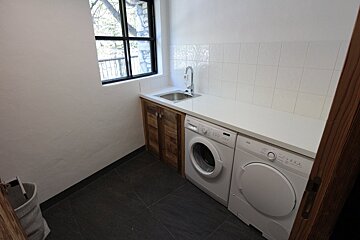 A modern laundry room features a white washer and dryer, a sink with white tiled backsplash, a light countertop, and wooden cabinets. A window overlooks trees, while dark floor tiles complete the spac