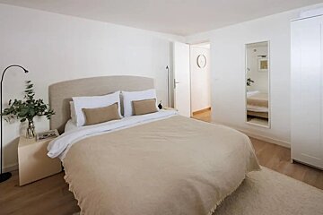 A minimalist bedroom with a large bed in neutral beige and white tones, light wood floors, and a full-length mirror.