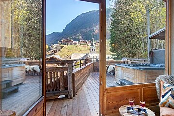 A wooden deck with hot tubs overlooks a scenic Alpine village and mountains, viewed through an open cabin door.