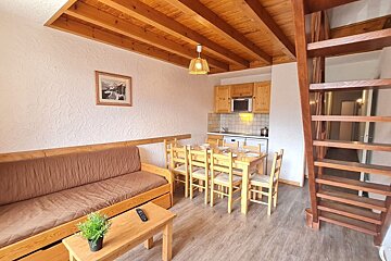 Cozy wooden apartment interior with a brown sofa, a dining table, a small kitchen, and an open wooden staircase.