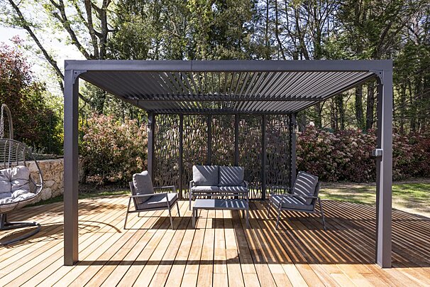 Modern gray pergola with matching patio furniture on a wooden deck, surrounded by a lush green garden.