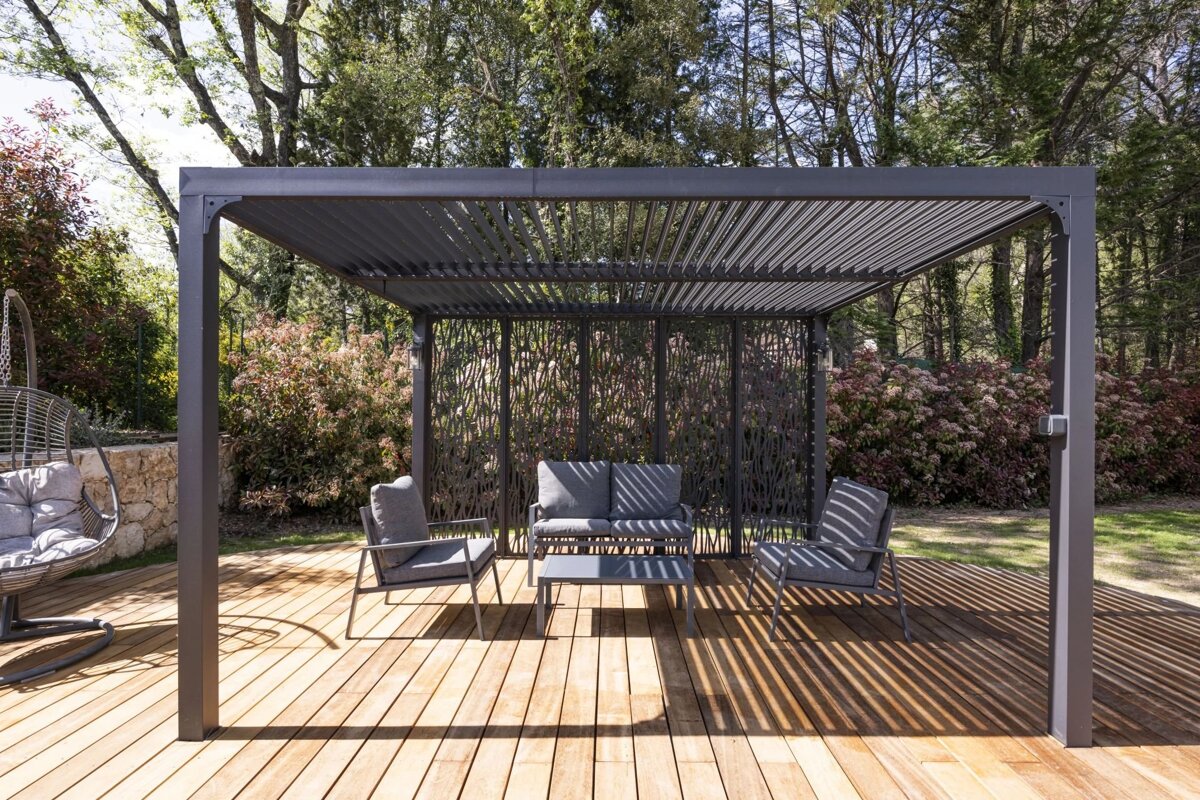 A gray pergola shades a wooden deck with outdoor seating, surrounded by green foliage. Sunlight casts striking striped shadows.
