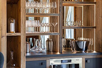 Modern wooden home bar featuring illuminated shelves filled with glassware, decanters, cocktail shakers, and an ice bucket.