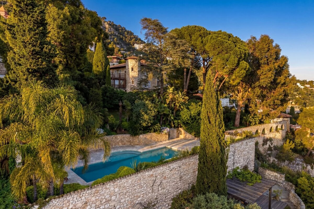 A scenic hillside estate featuring a swimming pool, stone walls, lush green trees, and a classic villa under a blue sky.
